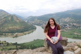 Girl standing on a high hill facing camera with a winding river, towns, and mountains in the background