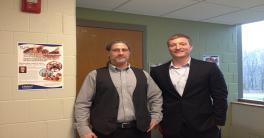 CSEES MA Alumnus Dusty Wilmes Two men standing side by side in front of a door and yellow wall